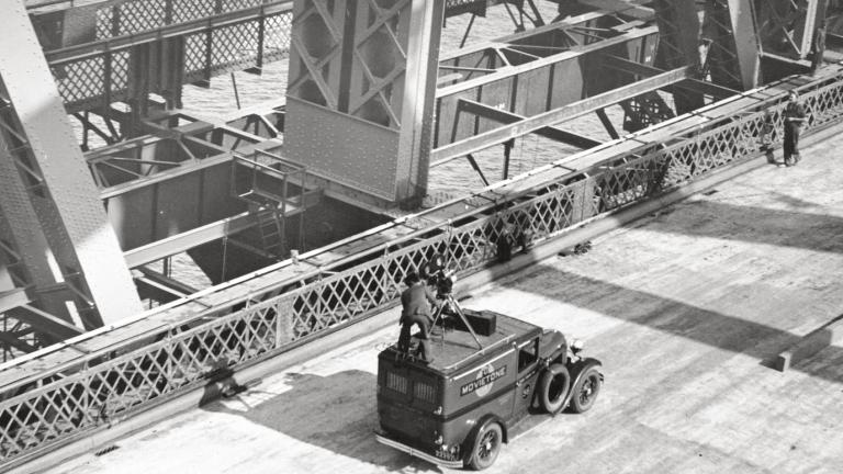 Fox Movietone Productions van with a man standing on top operating a camera. The van is on the deck of the newly-constructed Sydney Harbour Bridge.
