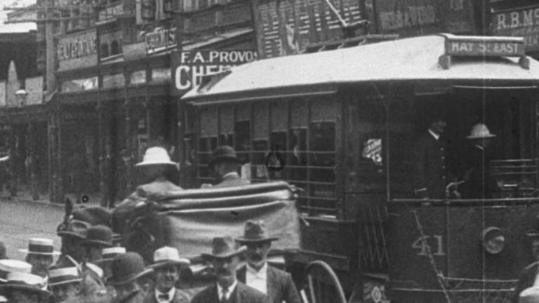 A tram goes past on a busy street in 1907. A horse and carriage is also going past. Men all wear hats. Shop hoardings promote businesses.