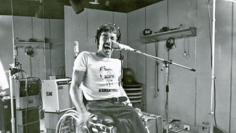 Singer Jeff St John in a recording studio, seated in a wheelchair, wearing headphones and singing with eyes closed into a microphone.