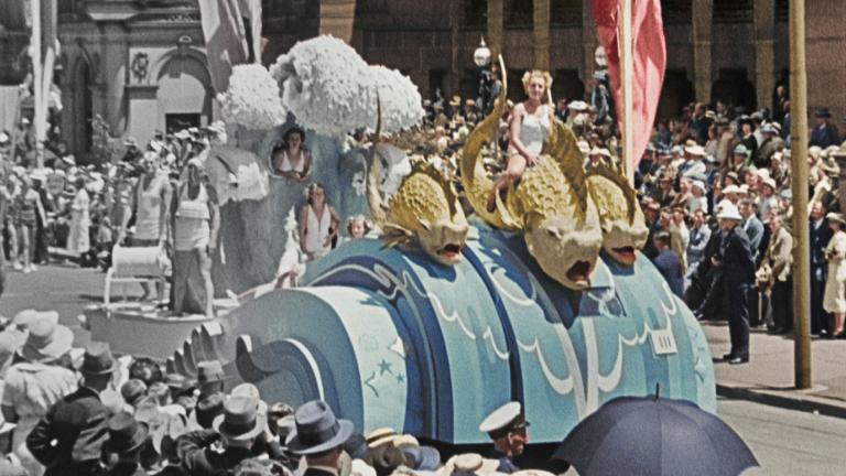 A partly colourised photo of a nautical-themed float in a 1930s street parade. On the float a woman dressed in white sits on a large-scale model of a sea creature.