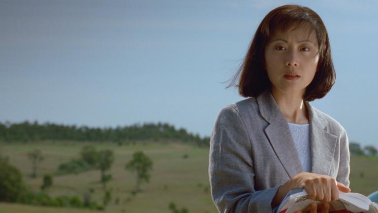 Still from NFSA Restores: Floating Life A young Asian woman dressed in a suit jacket pictured from the waist up in front of a green field and hills with trees in the background