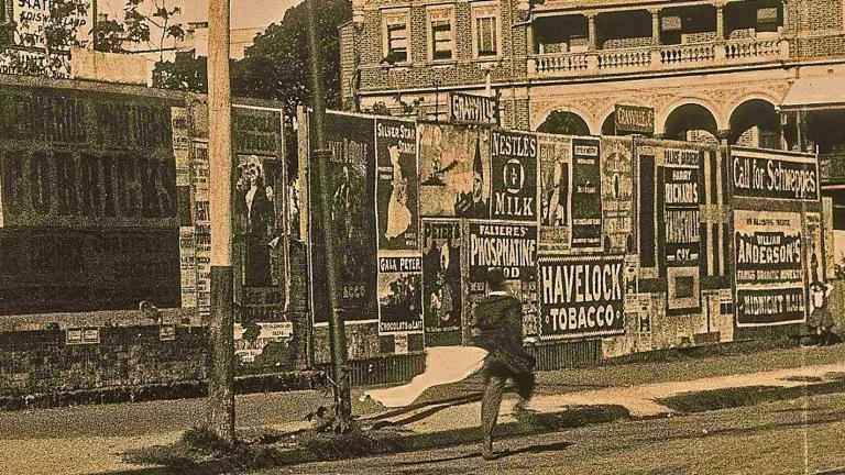 A scene from the early silent film Bashful Mr Brown shows a man walking down a street next to a fence lined with posters and advertisements.