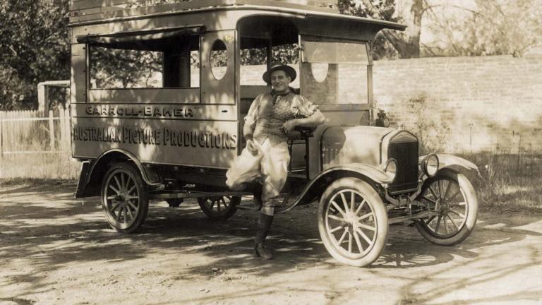 Actor and filmmaker Snowy Baker leaning against a car with 'Carroll-Baker Australian Picture Productions' written on the side