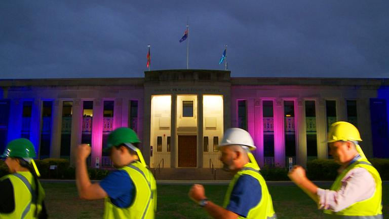 Dance Off_1600x775 Four people wearing protection helmets and hi viz shirst march in line in front of the NFSA building at night.