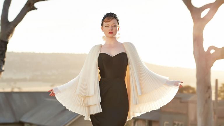 Saran Snook in black dress with white cape stands in front of an outback scene at sunset