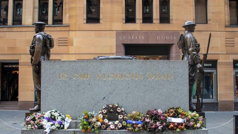 An Anzac Day memorial in Sydney inscribed 'To Our Glorious Dead' and flanked by statues of two soldiers. Wreaths of flowers surround the memorial