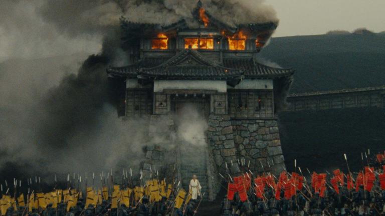 Film still from RAN of a Japanese castle on fire surrounded by an army.