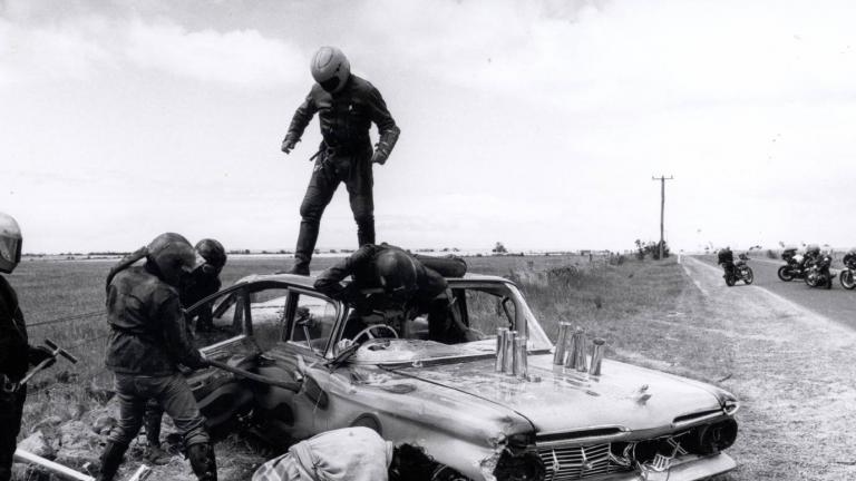 Members of a motorcycle gang smash up a car by the side of a highway