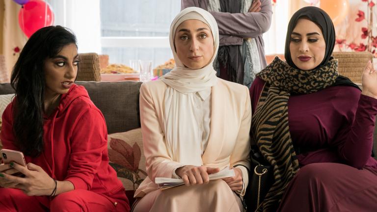 Three women sitting on a couch. One is looking sideways with an expression of annoyance at the other two women who are both looking straight ahead.