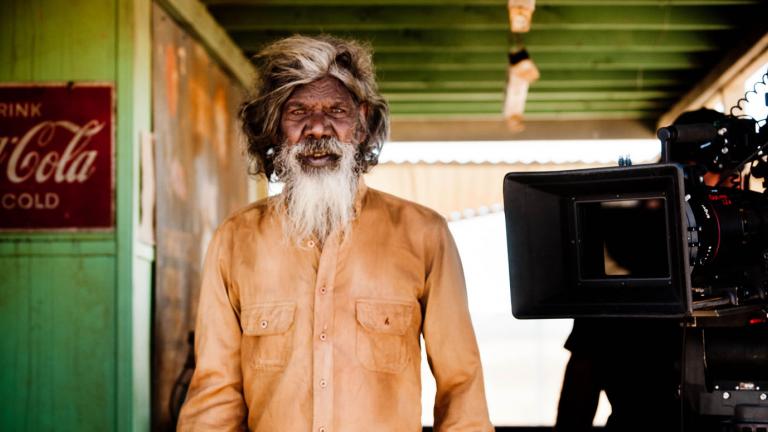David Gulpilil on the set of Satellite Boy with a film camera beside him. He is looking directly at the camera.