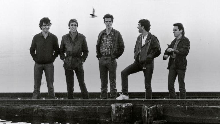 The five band members of Cold Chisel standing in a row on a pier with water in front and behind them. 