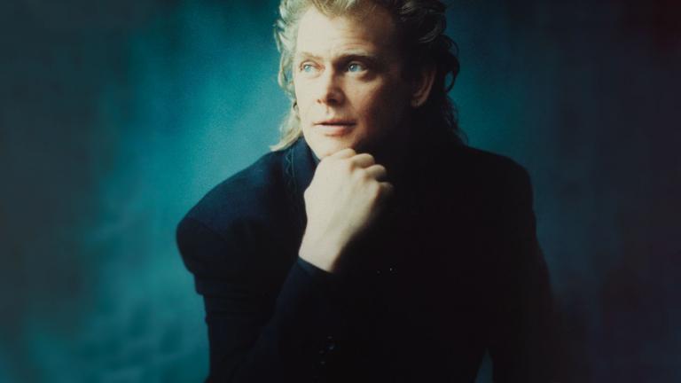 Image of John Farnham circa 1990 wearing a black jacket and seated with his chin resting in his hand.
