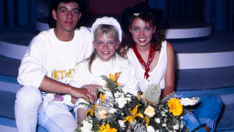 Young Talent Team members Vince Deltito, Rikki Arnot and Dannii Minogue sit together. Rikki, sitting in-between the two older teens, is holding a bunch of flowers.