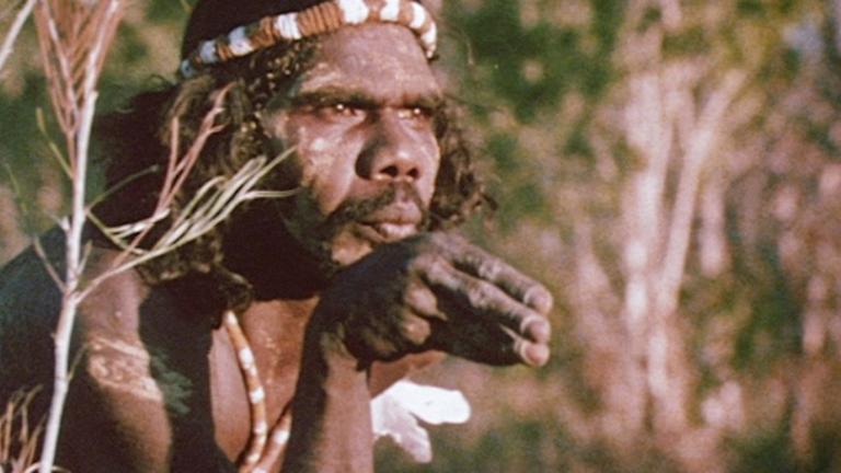 A young David Gulpilil is wearing traditional bodypaint in the bush. He is looking off-screen. He holds his hand in a dance pose in front of his mouth.