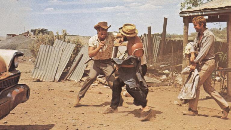 Jack Thompson and Donald Pleasence mock-brawling in the dusty outback while Gary Bond walks towards them