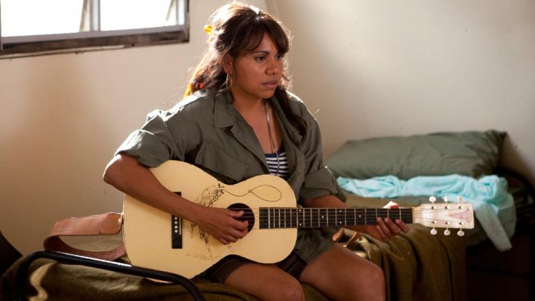 A woman sitting on a bed playing an acoustic guitar.
