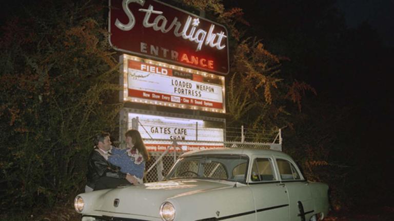 A man and woman embrace outside a drive-in cinema at night, leaning against their car