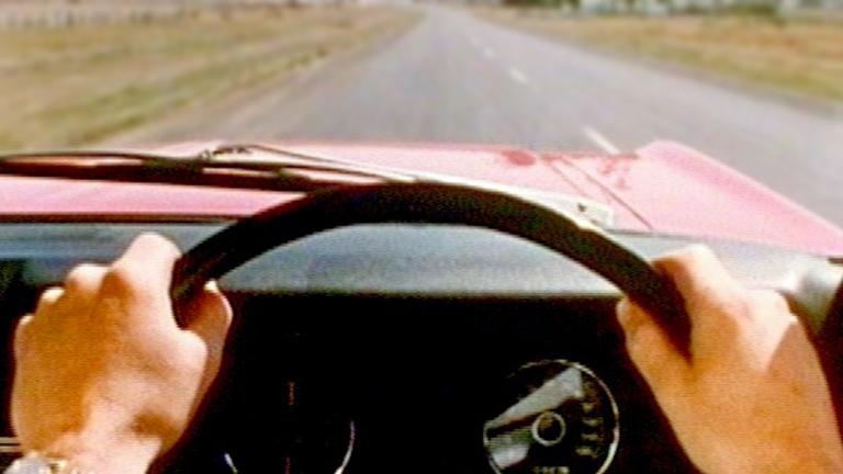A man is behind the wheel of a red XT Falcon. We see his point of view of the road ahead.