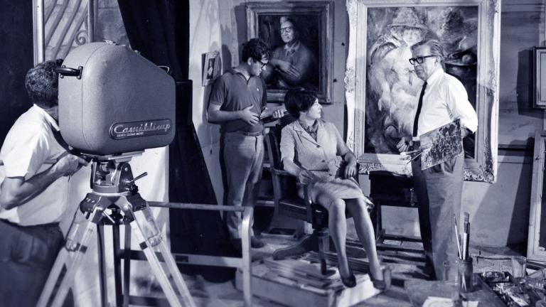 Blue-tinted black and white still of a man is operating a large film camera. He is filming artists Margaret Olley and William Dobell. There are two portraits painted by Dobell in the background as well as another member of the film crew.