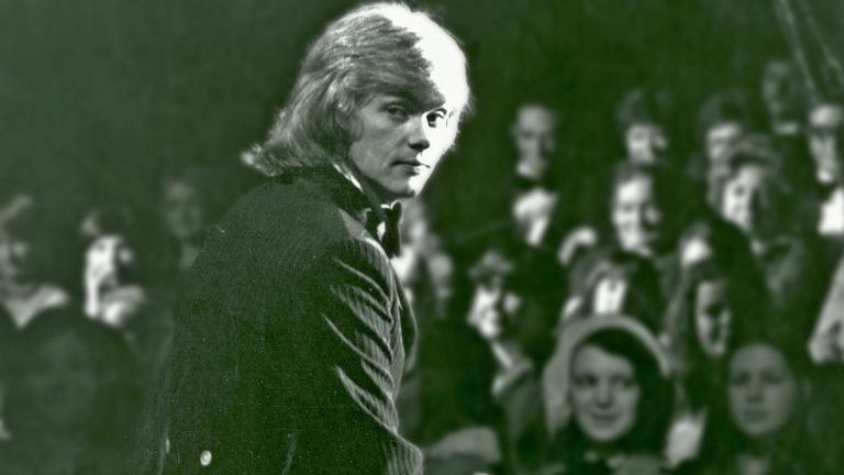 John Farnham sitting on stage in front of an audience, looking back over his right shoulder at the camer.