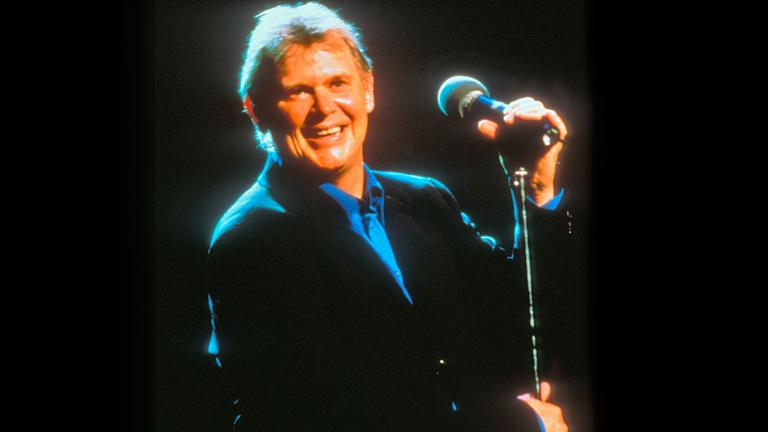 Singer John Farnham, circa late 1990s or early 2000s, pictured from the waist up wearing a black jacket and blue collared shirt, standing on a stage and holding on to a microphone stand. He's smiling at the camera.