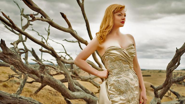 Actress Sarah Snook in a publicity shot for The Dressmaker, wearing a fitted, champagne-coloured, beaded gown and standing in front of some bare tree branches. In the distance you can see a brown, dusty plain and grey, cloudy sky.