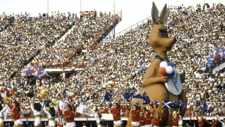A packed stadium of people witnessing the opening ceremony of the Commonwealth Games. Matilda the huge mascot of the games is on the field along with lots of school children.