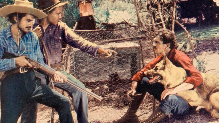 Two men hold rifles and stand over a man sitting on the ground with a dog