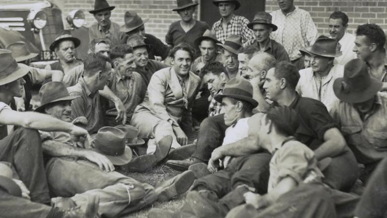 Actor Charles Farrell reclines on the ground surrounded by a crowd of men.