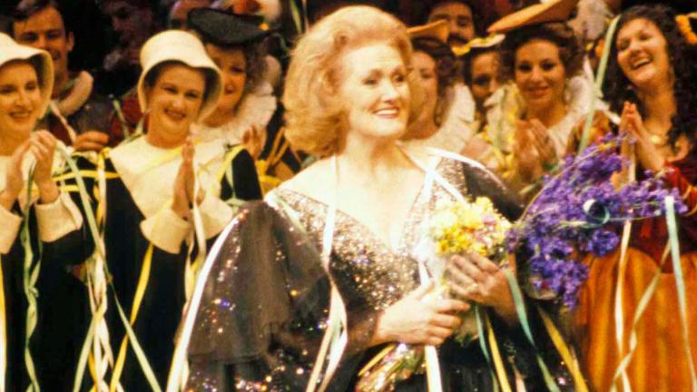 Dame Joan Sutherland on stage holding flowers with the cast applauding her.