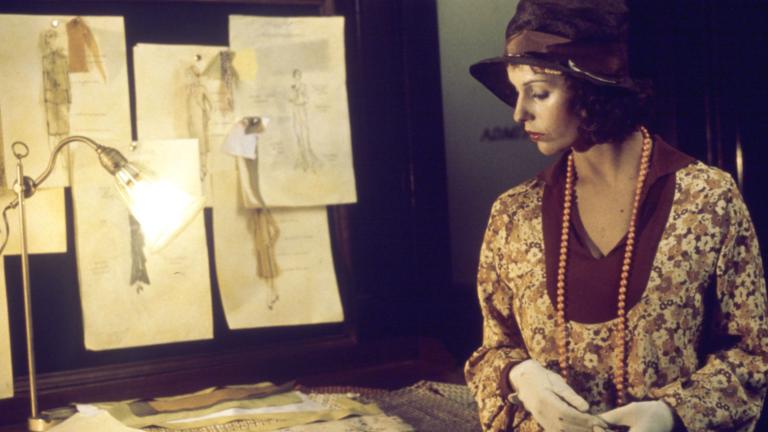 Film still of Helen Morse as Caddie looking at drawings of women's dress designs in a studio.