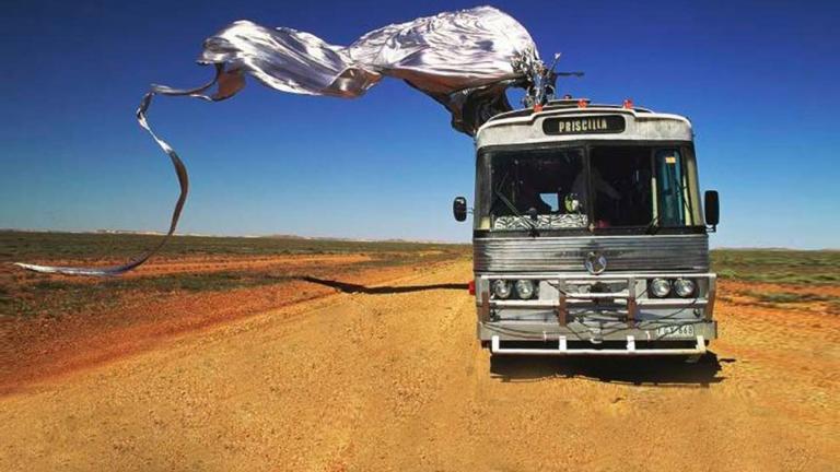 Scene from Priscilla, Queen of the Desert. A tour bus on a dirt road in the desert with blue sky in the background. On top of the bus is Guy Pearce in a shiny silver frock sitting on a giant stiletto shoe with a long train flowing behind the bus.