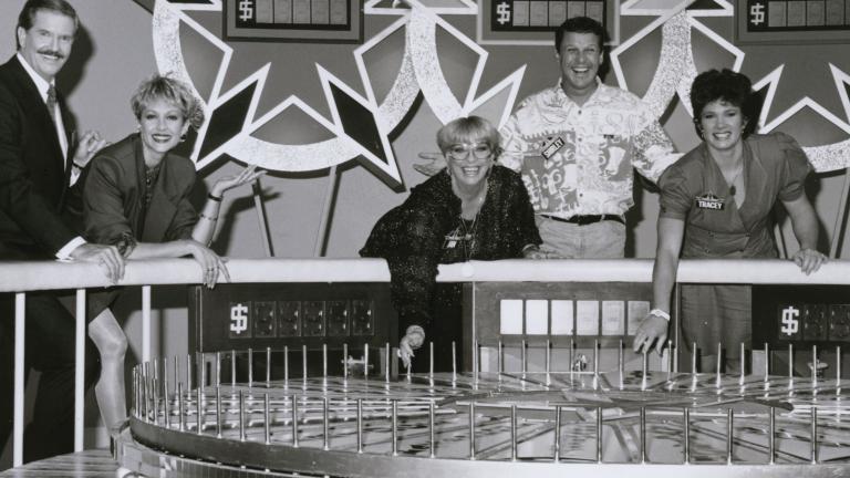 Game show hosts and contestants in a TV studio standing behind a big spinning wheel.