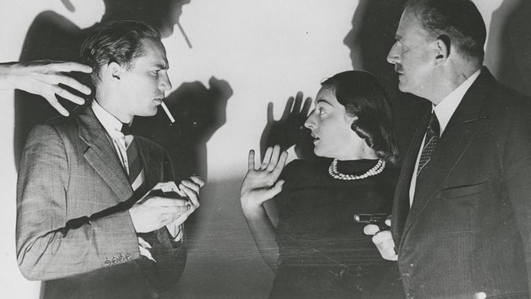 Two men and a woman. One man lights a cigarette while a mysterious hand reaches for him. The woman looks terrified. The other man holds a gun.