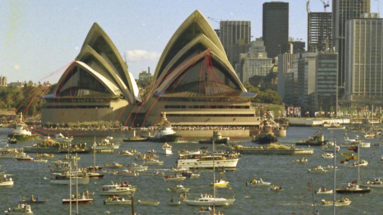 The Sydney Opera House is festooned with red ribbons. There are many boats on the water in front of it.