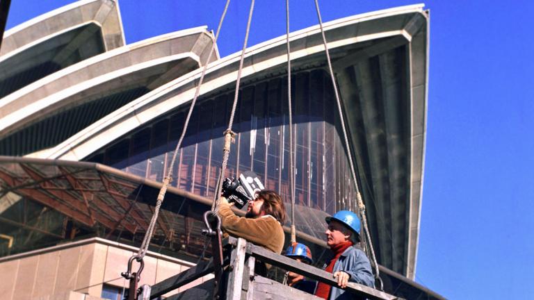 The sails of the Opera House tower above a camera operator and a man in a hardhat. They are standing in a wooden box which is hoisted to a crane.