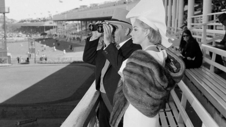 A man and woman are dressed for a day at the races. He is looking through binoculars. Black and white image.