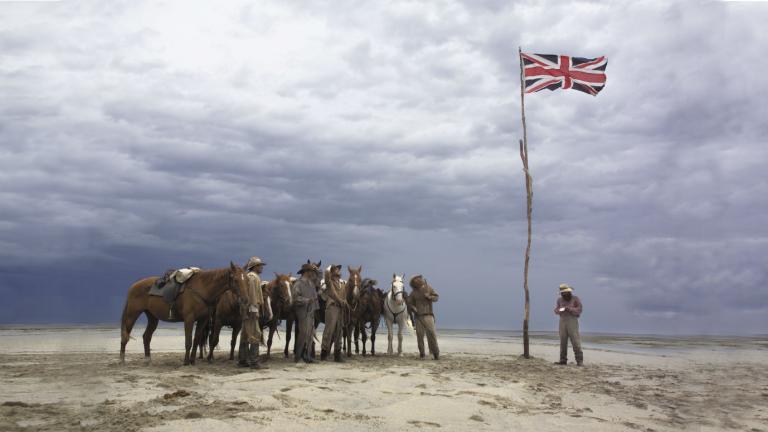 A group of men and horses standing on a plain in the Australian outback stand looking at a Union Jack flag that has been raised on a long, thin tree trunk.