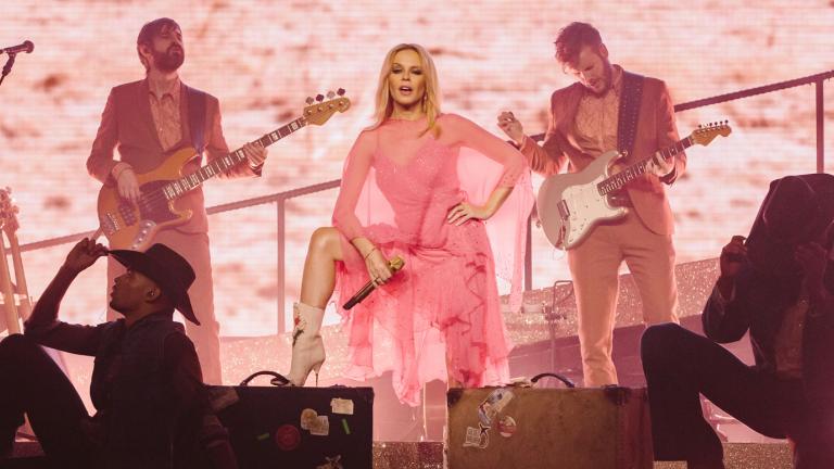 Kylie Minogue is standing on a stage with a foot up on an amplifier in a power pose. There are dancers in front of her and two musicians on stage with her playing guitar.