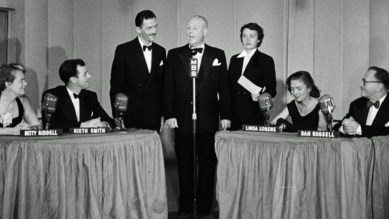 A studio with 3 people standing in the centre in front of a radio microphone and a table on each side with two panellists seated at each table.