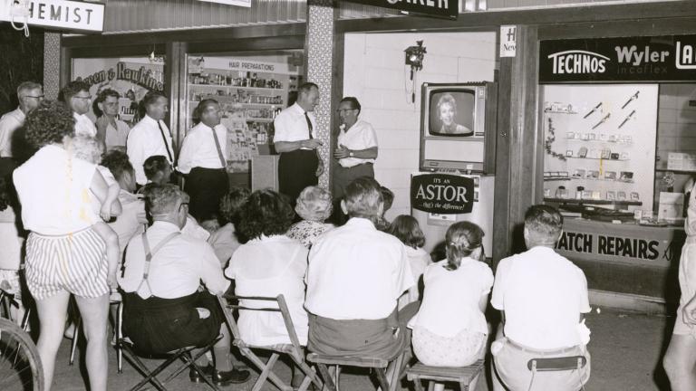 A crowd of people (some seated and some standing). They are watching a television set through a shop window.