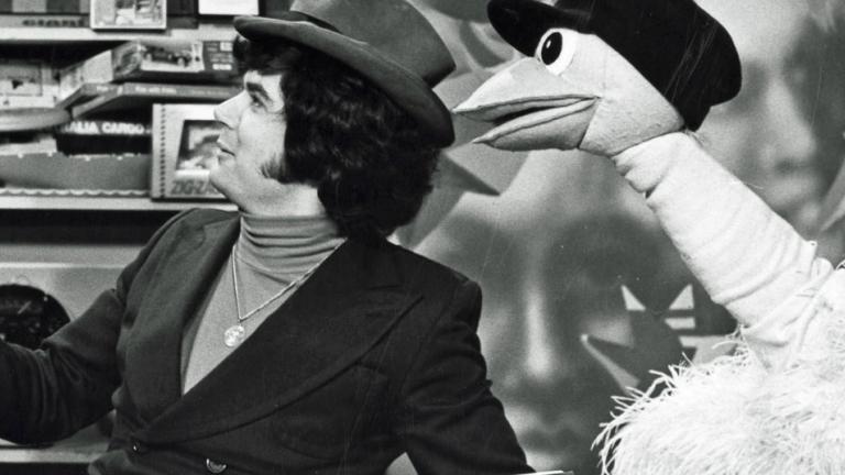 A man wearing a hat sitting next to an ostrich puppet who is also wearing a hat. They are both turned to their right.