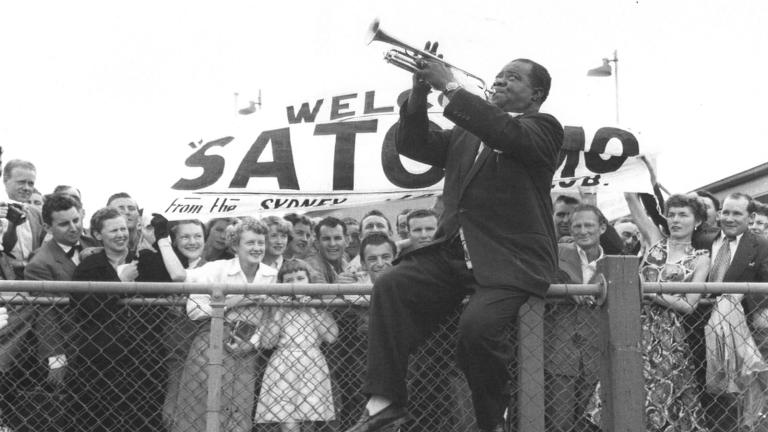 Louis Armstrong at Sydney airport in 1956, He's sitting on the top rail of a wire fence, playing the trumple to a crow gathered there to welcome him.
