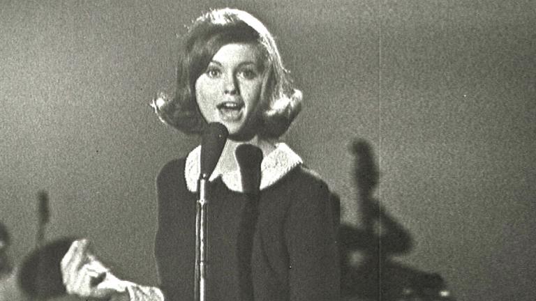Olivia Newton-John at age 16 in a TV studio standing behind a microphone singing.