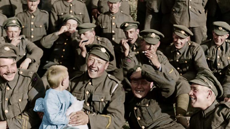 Three rows of British soldiers in uniform relaxing on the grass. One at the front is smiling and holding a small child.