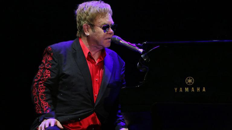 Musician Elton John sitting at a piano and singing into a microphone. He is wearing a black and red suit and dark glasses. 