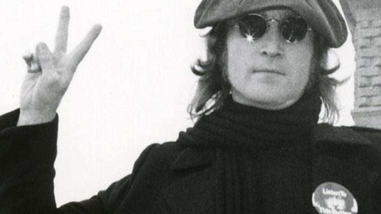 Head and shoulders shot of John Lennon pictured in New York, wearing sunglasses and a beret and making a peace sign with his fingers. C. 1973.