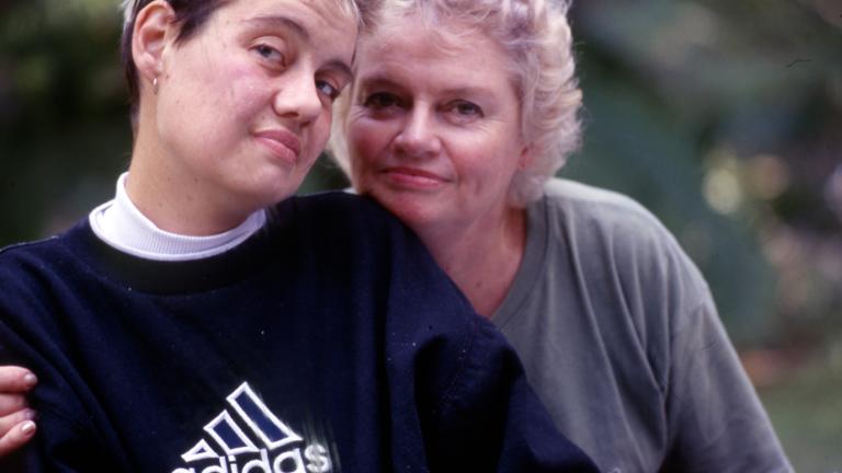 A young woman with short blond hair, wearing a black jumper is embraced by an older blond lady. They are looking at the camera.