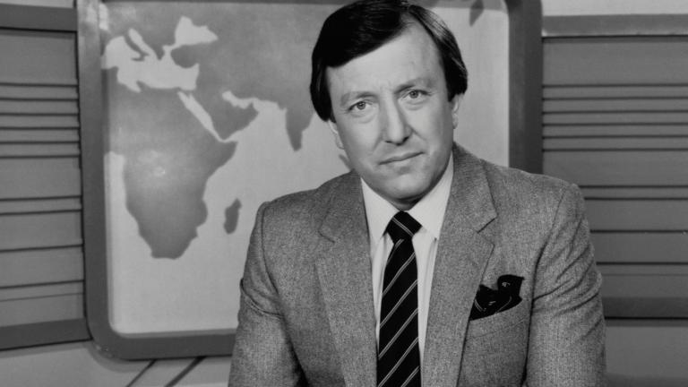 Journalist Jim Waley is wearing a suit and tie in this black and white photo. He is sitting in a studio with a backdrop showing a map of the world.