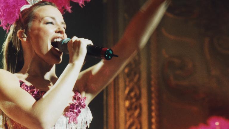 Kylie Minogue on stage in a pink showgirl costume singing into a microphone.